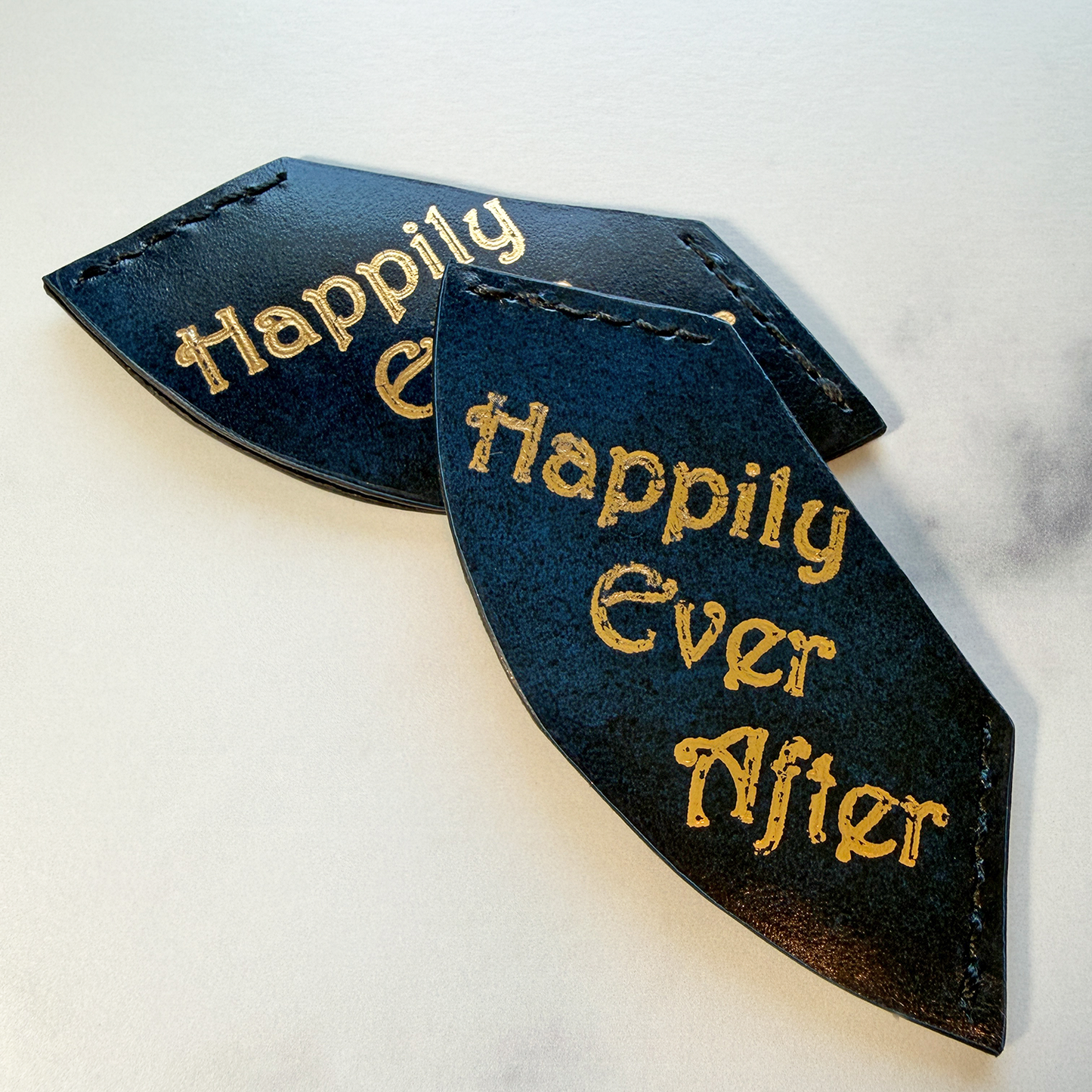Two blue & black leather corner bookmarks with 'Happily Ever After' gold foil text on a marble countertop