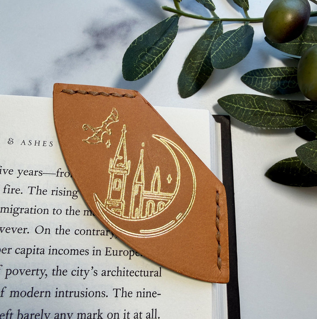 A tan calfskin leather corner bookmark with a gold foil picture of a small dragon flying over a castle, sitting inside of a crescent moon.