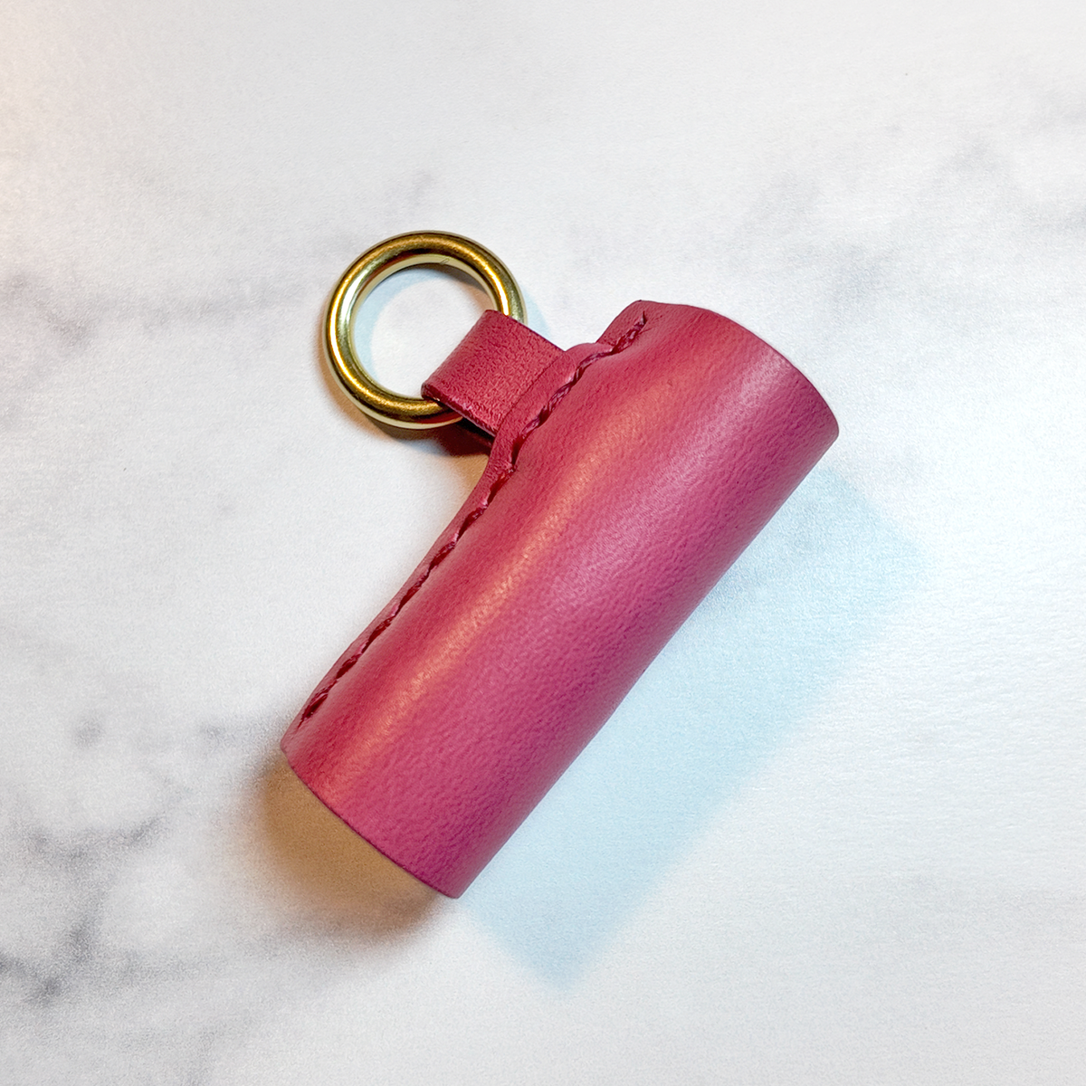 A pink leather cylindrical lip balm holder with a brass metal key ring, resting on a white marble surface.