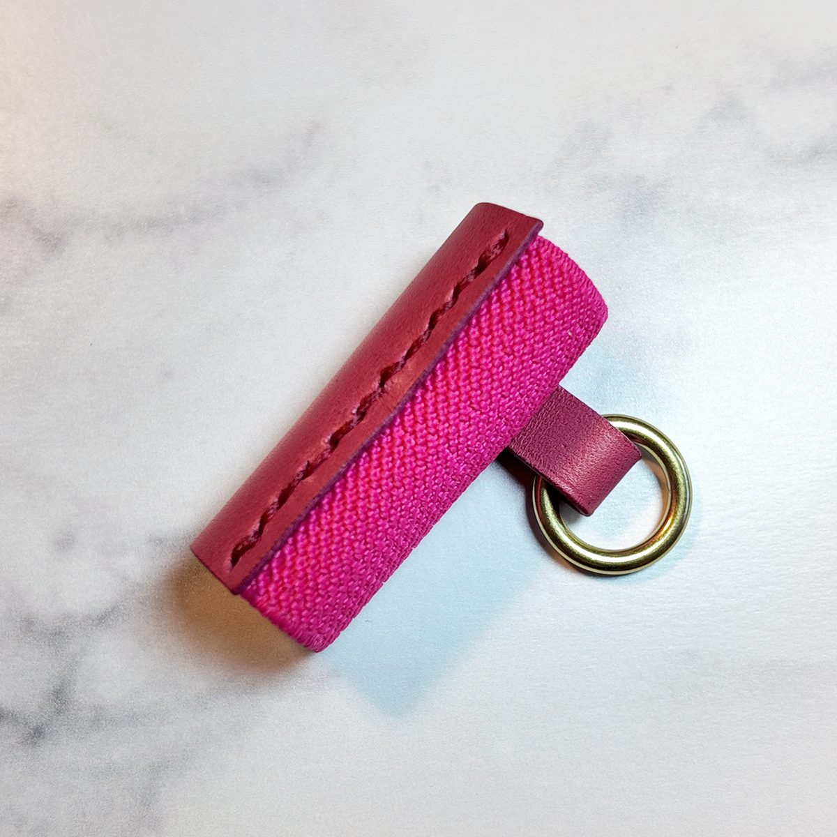 A hot pink leather and elastic lip balm holder with a brass metal key ring, resting on a white marble surface.