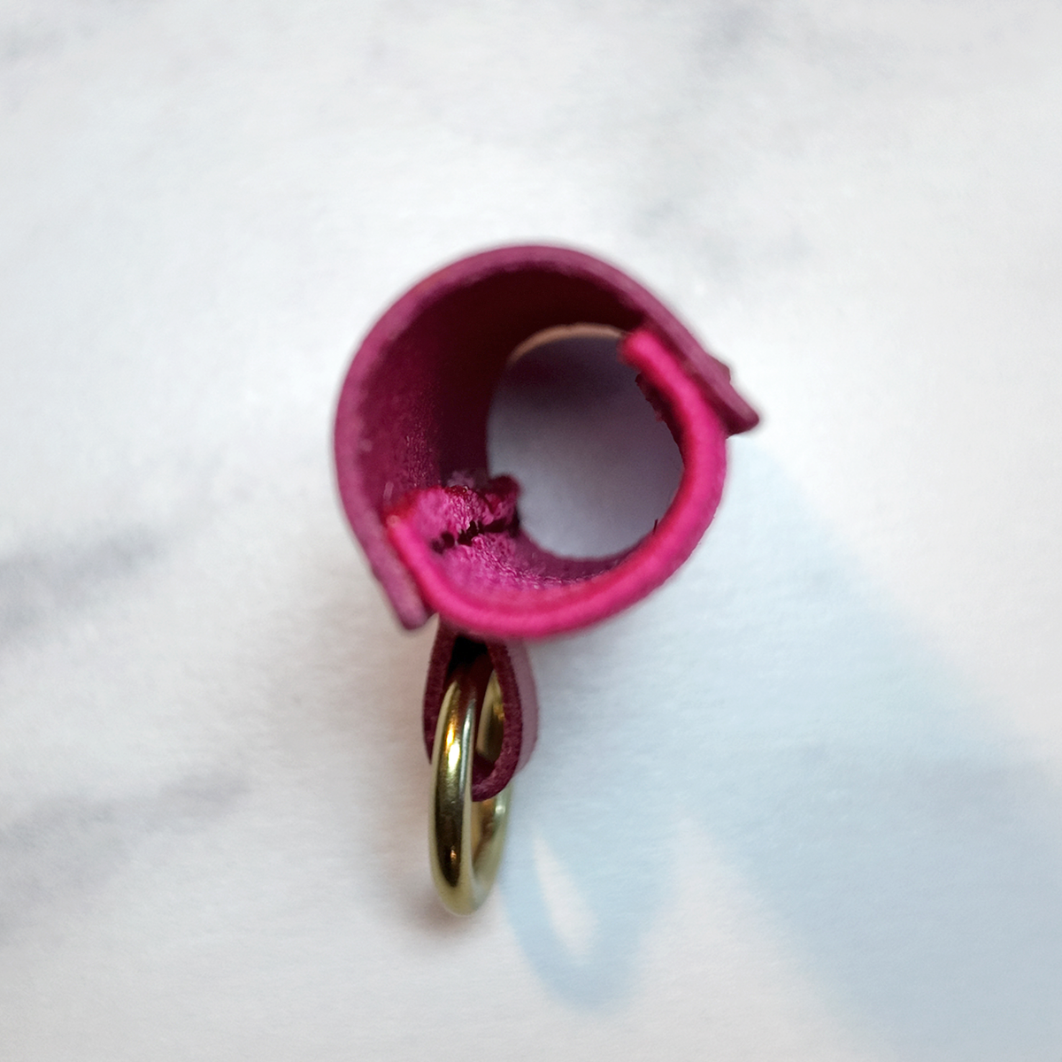 A close-up of a hot pink leather lip balm holder with a brass metal key ring, resting on a white marble surface.