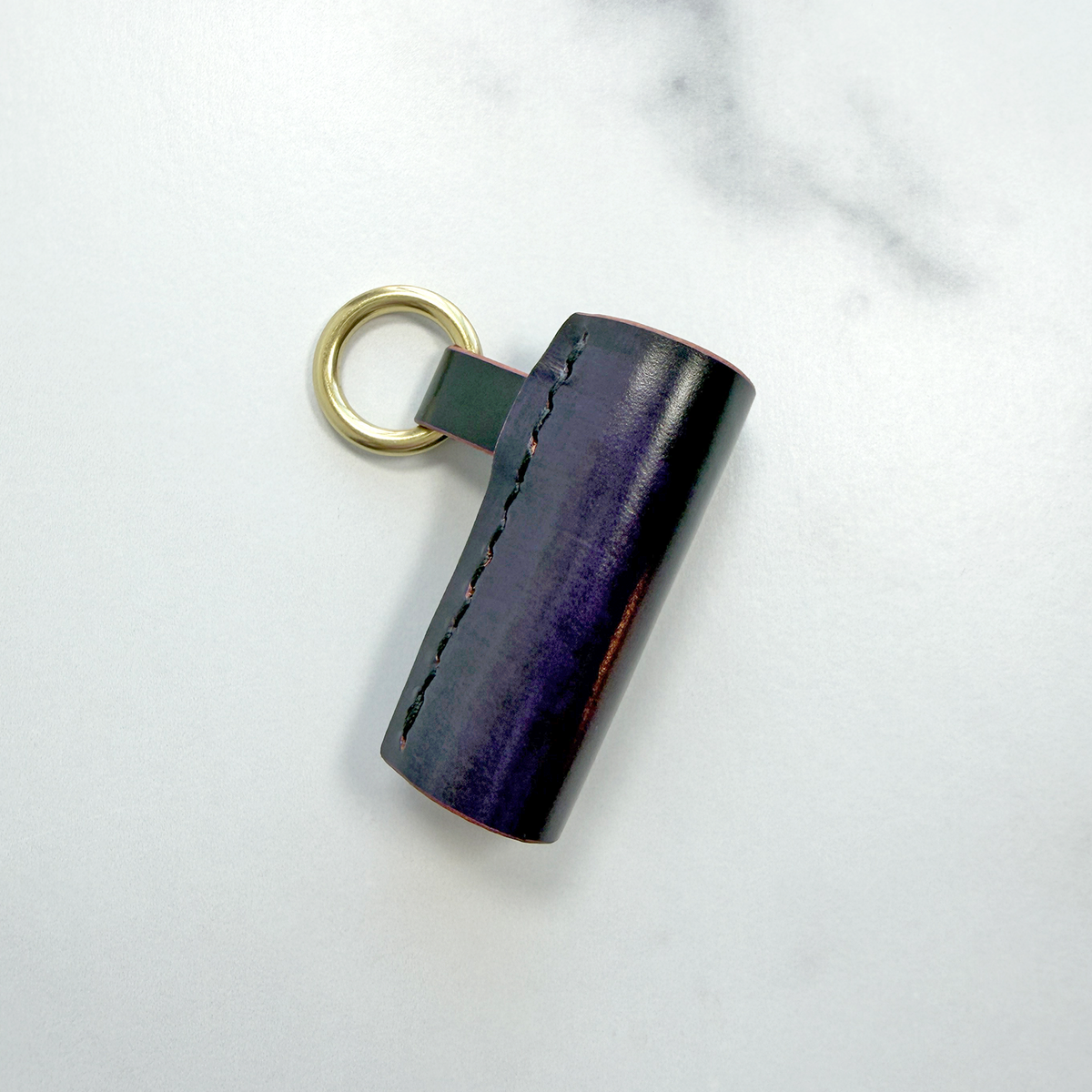 A purple leather lip balm holder with a brass ring to attach to a key chain on a white marble background.