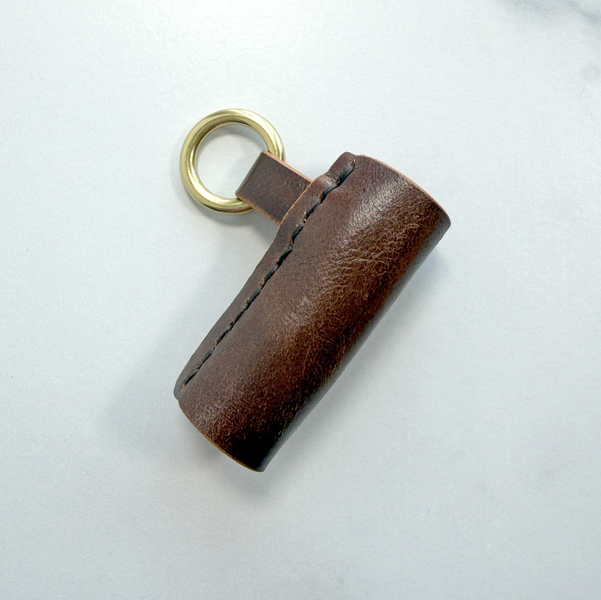 A brown leather lip balm holder with a brass ring to attach to a key chain on a white background.