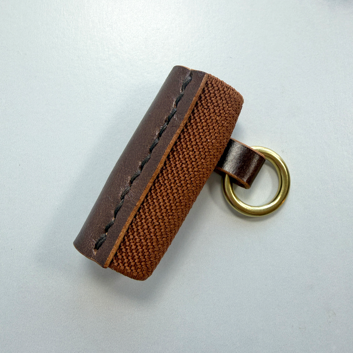 A brown leather lip balm holder with an elastic back, and a brass ring to attach to a key chain on a white background.