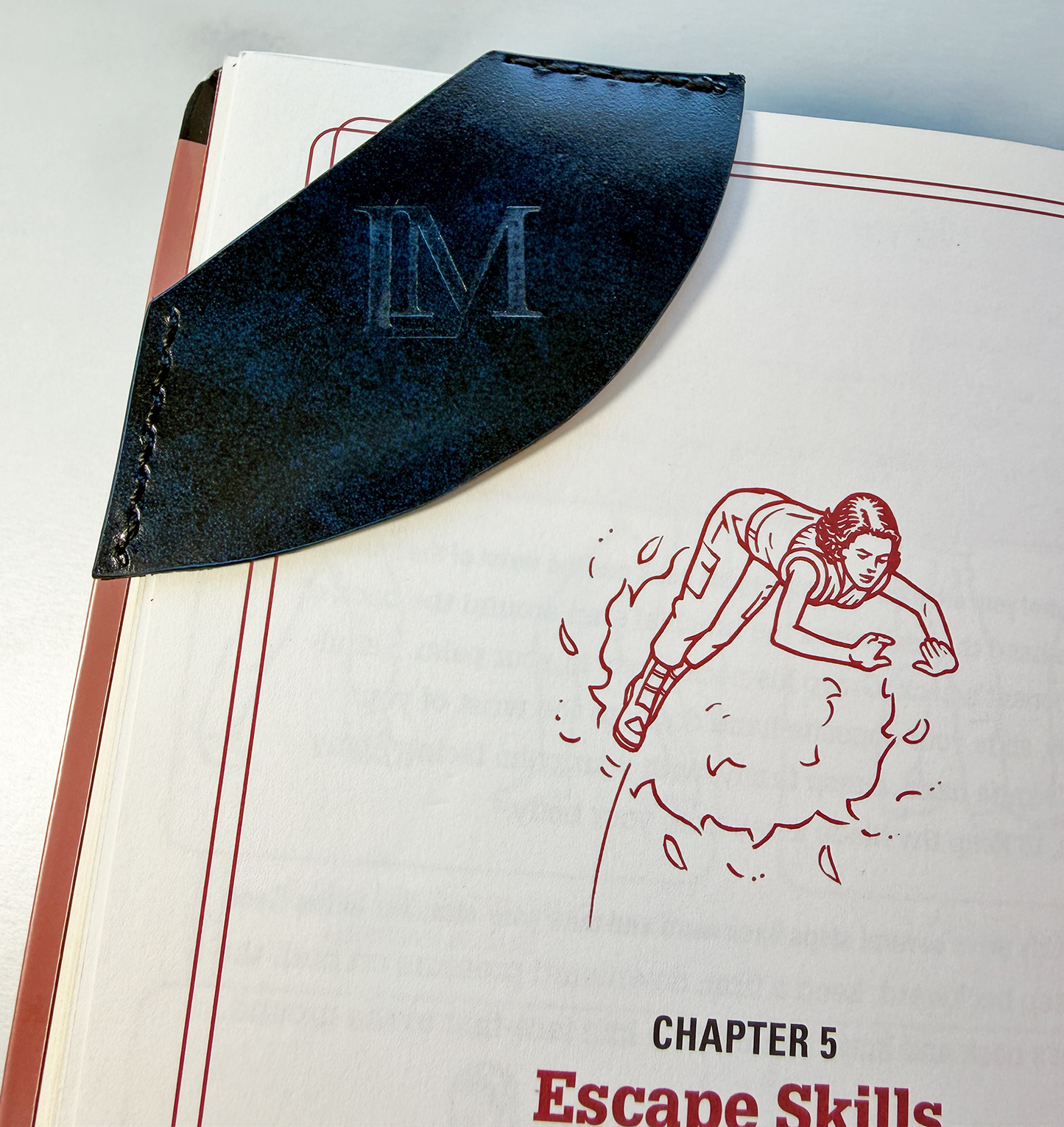 Blue& black leather corner bookmark with LM logo on a book chapter titled 'Escape Skills'.