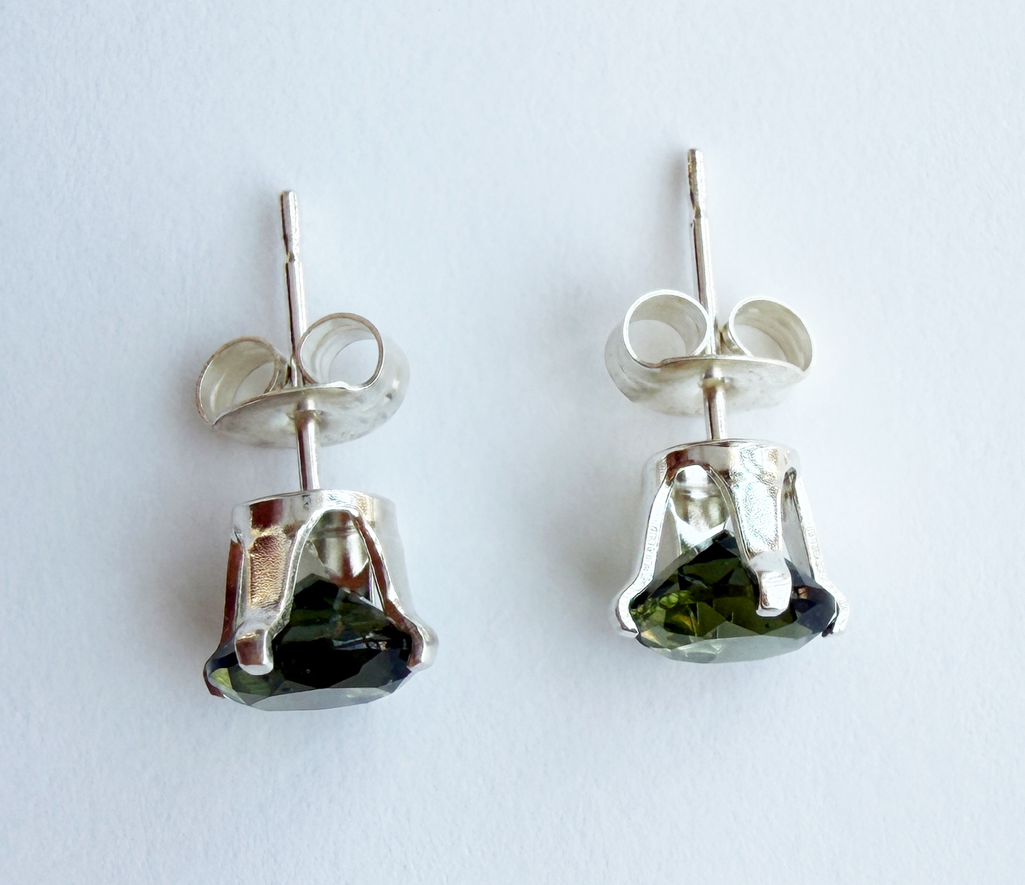 A side view of a pair of silver post earrings with green Moldavite gemstones on a white background
