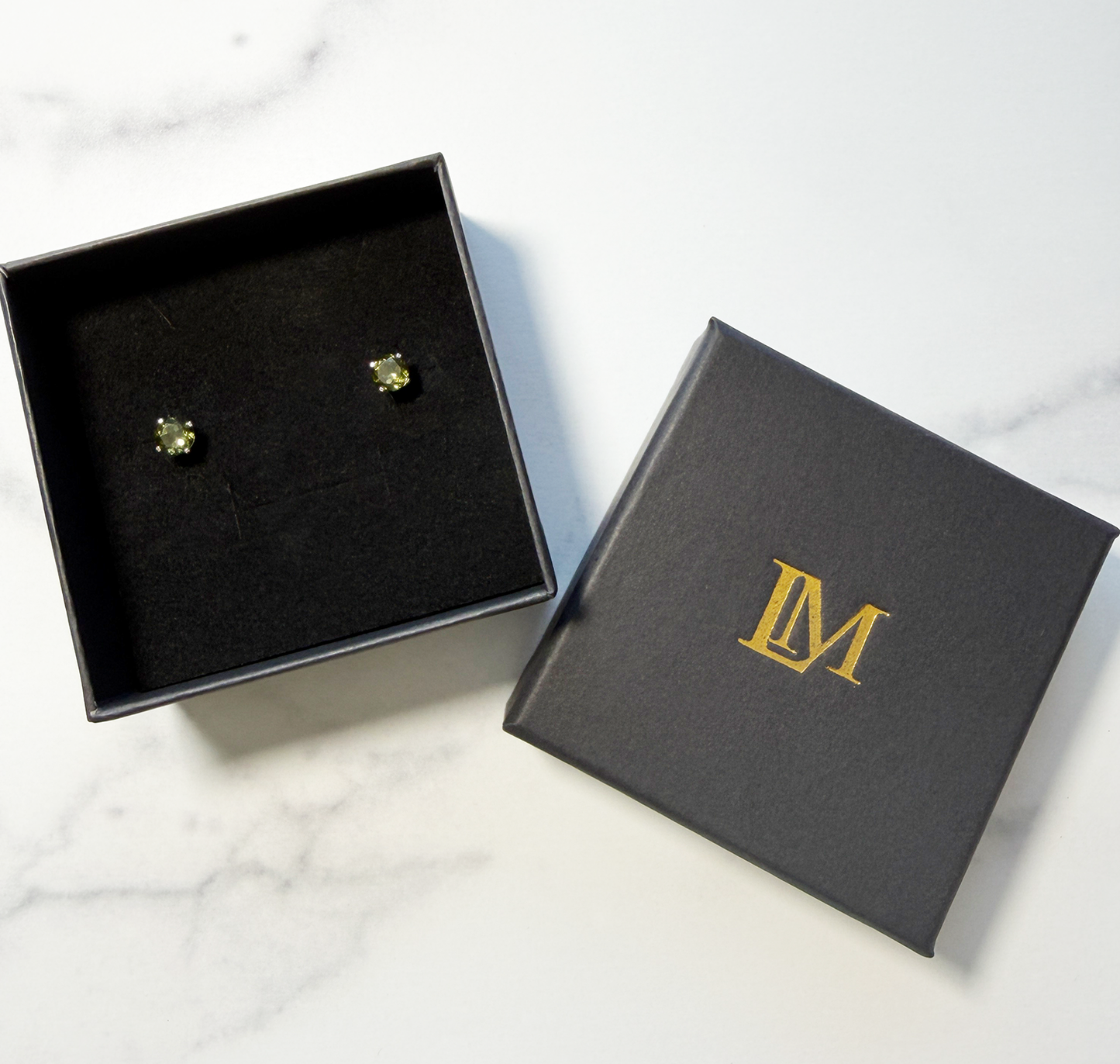 Jewelry box with 'LM' logo and a pair of green Moldavite gemstone stud earrings inside, on top of a marble surface