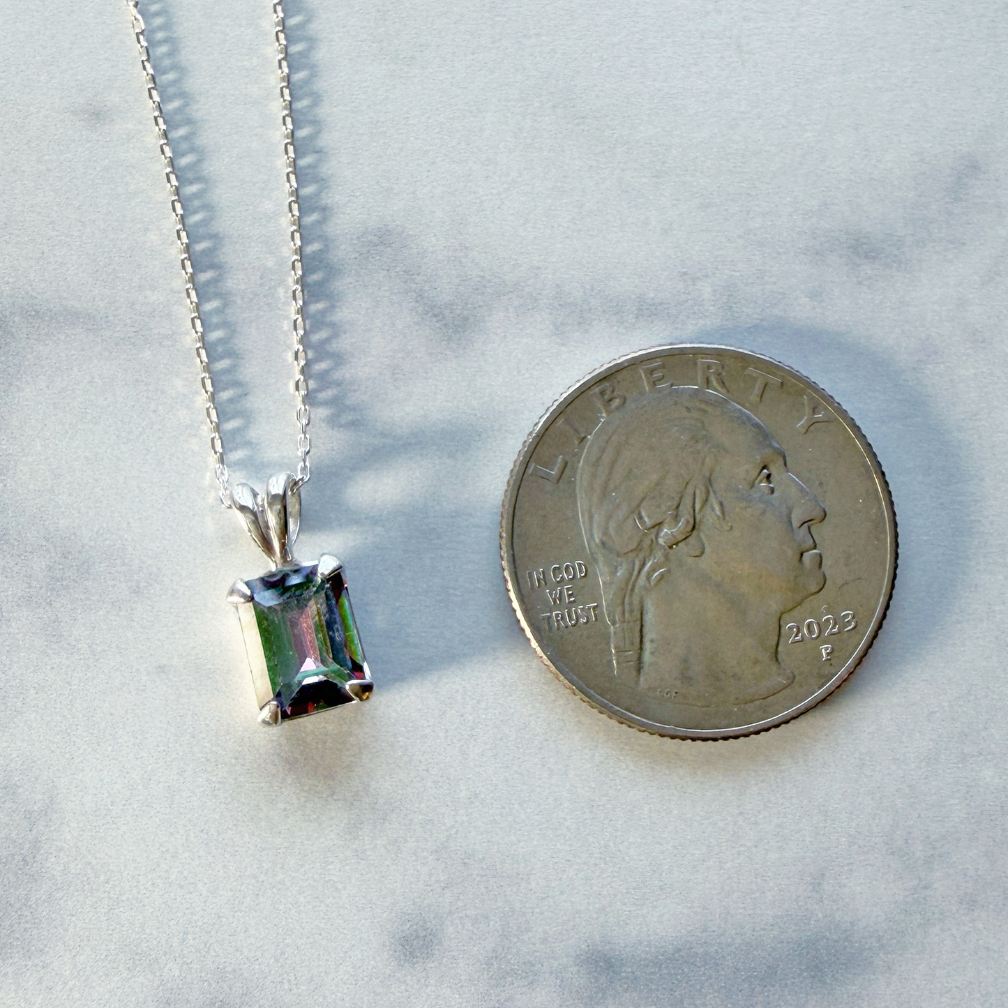 Sterling silver necklace with a mystic quartz gemstone pendant next to a quarter for scale on a marble surface