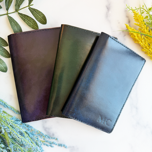 Three leather passport covers on a marble countertop. The passport covers are purple, green, and blue. The blue cover has engraved initials in the bottom right corner. 
