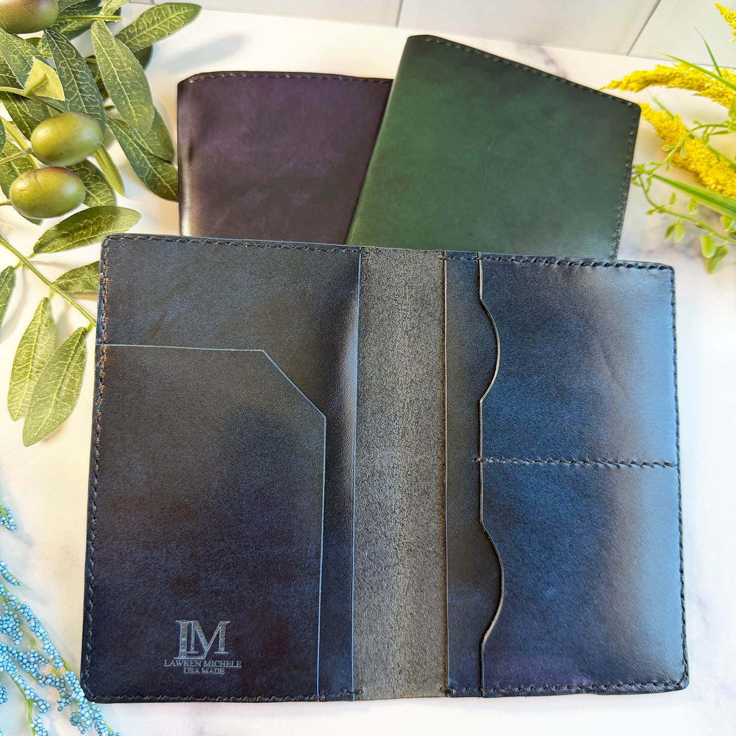 Three leather passport covers on a marble countertop. The passport covers are purple, green, and blue. The blue cover is open to show card & ticket compartments.  It also has a small engraved logo in the inside. 